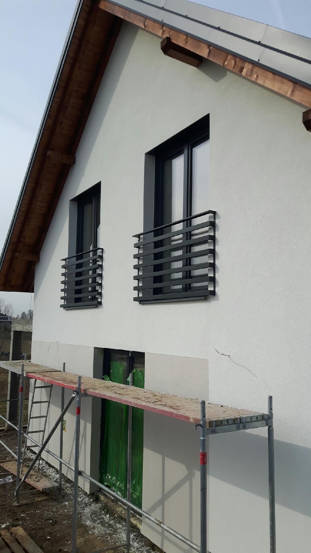 Nowoczesny dom z białym tynkiem i czarnymi, poziomymi balustradami okiennymi, widoczne rusztowanie i okno zabezpieczone folią ochronną.