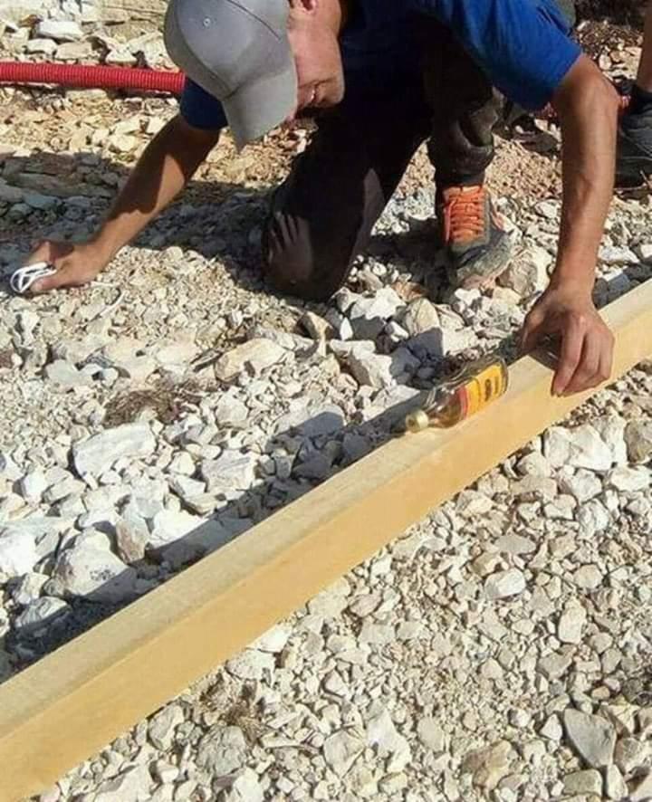 Mężczyzna w czapce klęczy obok drewnianej belki, używając butelki jako poziomicy na gruncie z kamieni.