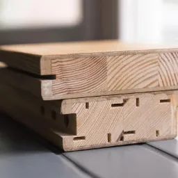 Przekrój przez drewniane elementy konstrukcyjne z widocznymi otworami i warstwami klejonego drewna, ułożone na szarym blacie przy oknie.