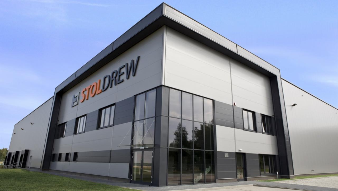 Nowoczesny budynek magazynowy z szaro-białą elewacją i dużymi oknami, logo firmy 'STOLDREW' w kolorze pomarańczowo-czarnym na fasadzie, zielony trawnik przed budynkiem, błękitne niebo w tle.
