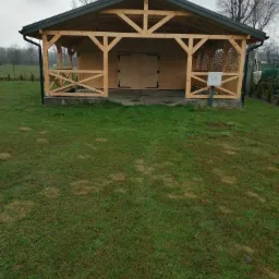 Drewniana altana z kratką na bocznych ścianach, z dwuskrzydłowymi drzwiami, na betonowej wylewce, otoczona trawnikiem i drzewami.