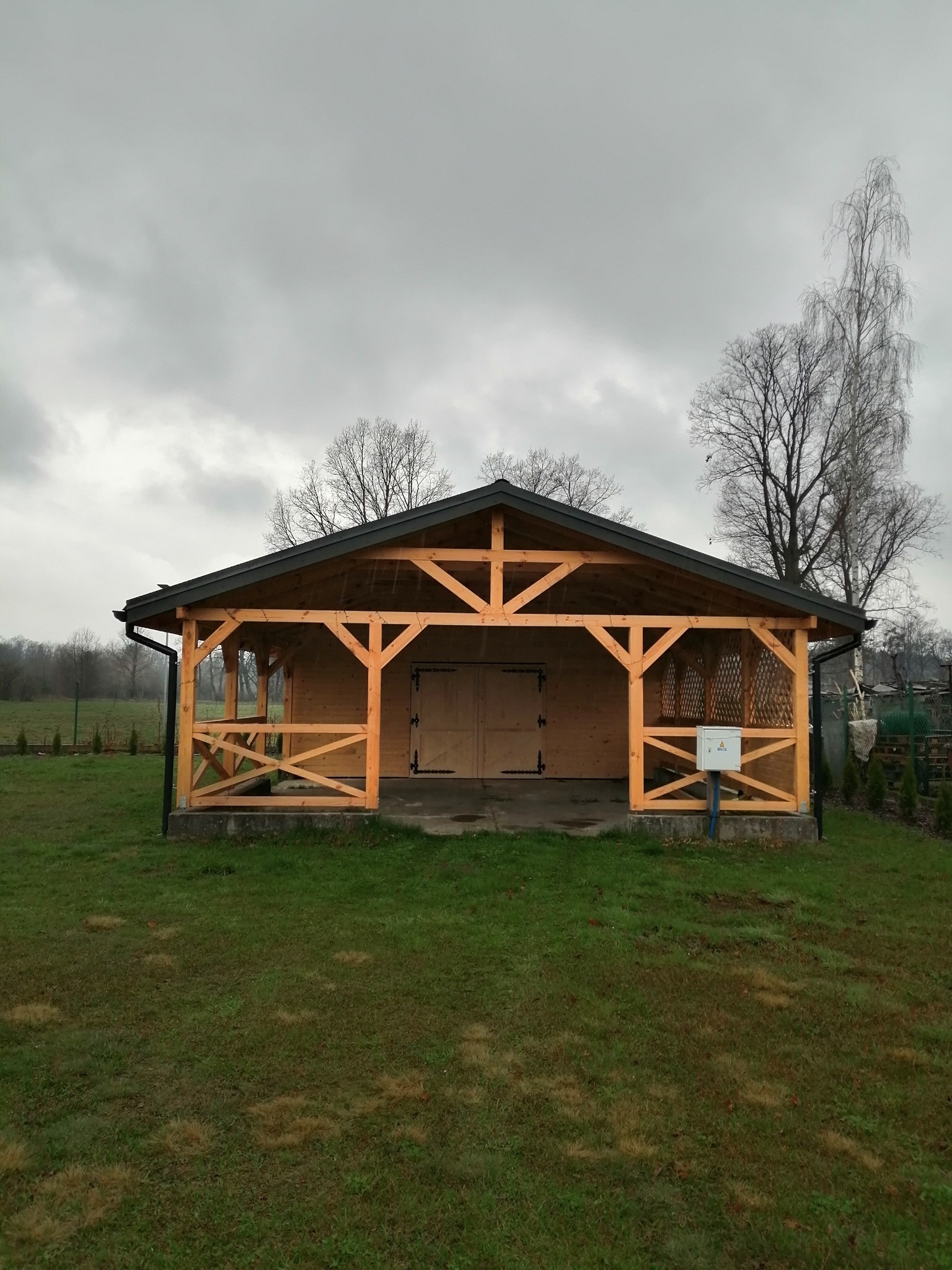 Nowa, drewniana altana z dwuspadowym dachem i ażurowymi ścianami, umiejscowiona na trawniku w pochmurny dzień.