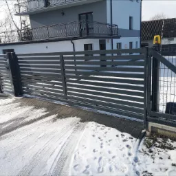Nowoczesna, pozioma brama wjazdowa z automatyką i lampą ostrzegawczą, połączona z siatką ogrodzeniową, na posesji pokrytej śniegiem.