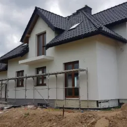 Nowoczesny dom jednorodzinny z białą elewacją, ciemną dachówką i drewnianymi oknami, w trakcie prac wykończeniowych, widoczne rusztowania i niezagospodarowany teren wokół budynku.