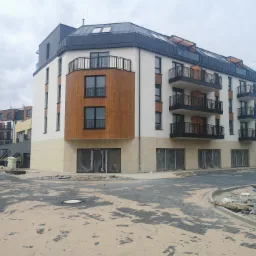 Nowoczesny budynek wielorodzinny z białymi i drewnianymi elementami elewacji, czarnymi balkonami i dachem, w trakcie wykańczania terenu wokół budynku.