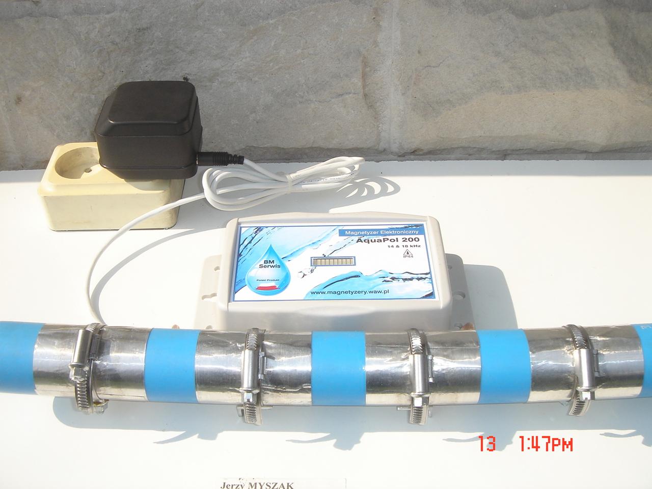 Elektroniczny magnetyzer wody AquaPol 200 zamontowany na rurze wodociągowej z niebieskimi paskami, zasilacz podłączony do gniazdka elektrycznego, widoczne logo BM Serwis.