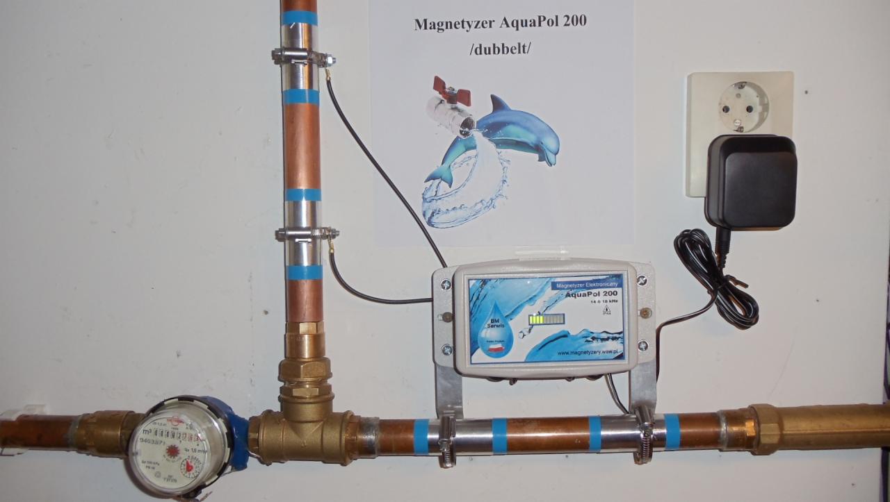Instalacja z licznikiem wody, rurami miedzianymi z niebieską taśmą, podłączonym elektronicznym magnetyzerem AquaPol 200 i zasilaczem sieciowym, na tle białej ściany z plakatem reklamowym...