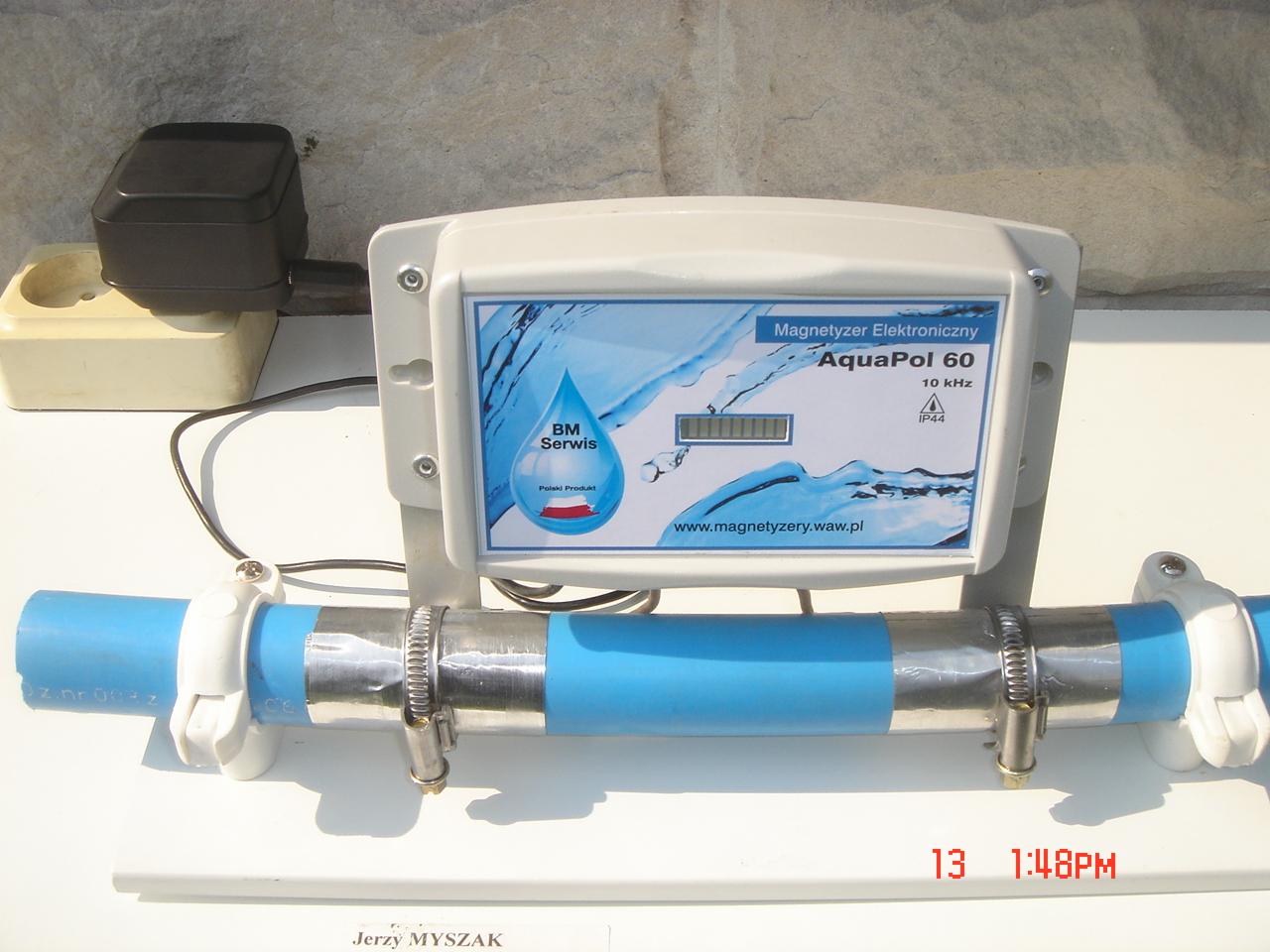 Elektroniczny magnetyzer wody AquaPol 60, zamontowany na niebieskiej rurze z metalowymi obejmami, z widocznym panelem sterowania i adresem strony internetowej producenta