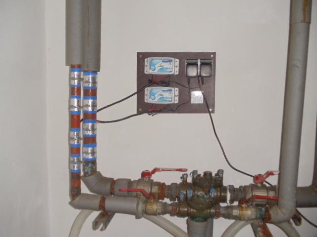 Instalacja wodna z rurami owiniętymi taśmą, podłączona do dwóch elektronicznych liczników na brązowej płycie, z widocznymi kablami i zaworami.
