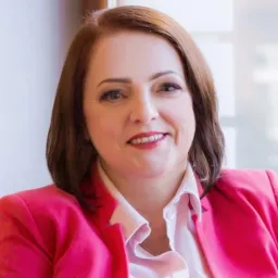 Portret kobiety w różowej marynarce i białej koszuli, uśmiechniętej, z krótkimi brązowymi włosami, na jasnym tle, w naturalnym świetle.