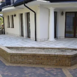 Taras z jasnoszarej kostki ułożonej we wzór, obrzeże z kostki w odcieniach brązu, donica z cegły, biały budynek z kolumną w tle.