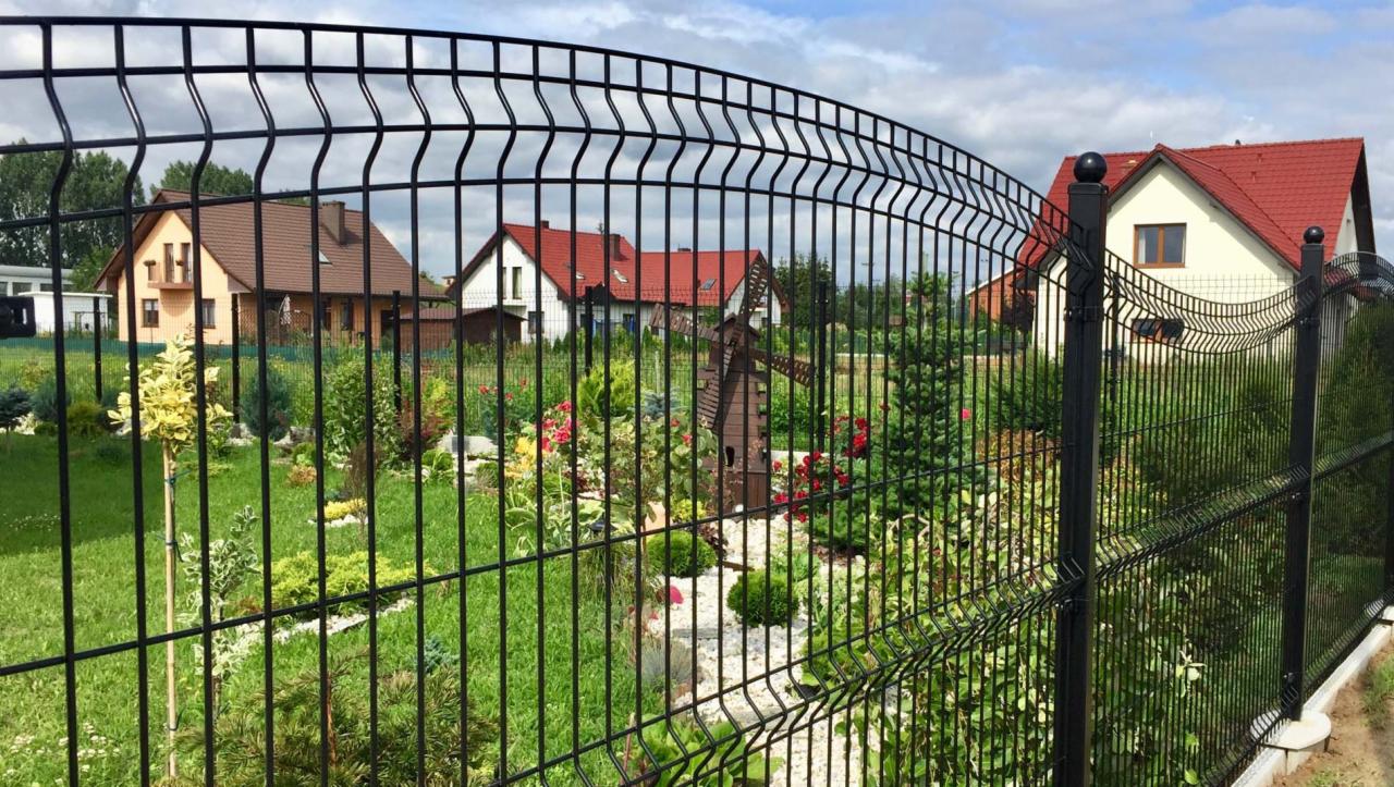 Czarne ogrodzenie panelowe 3D typu A z widocznym ogrodem, domami i drewnianym wiatrakiem w tle.