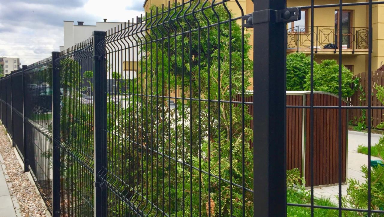Czarne ogrodzenie panelowe 3D z widocznymi zgrzewami, montowane w Zielonej Górze, z zielonymi krzewami w tle i fragmentem żółtego budynku.