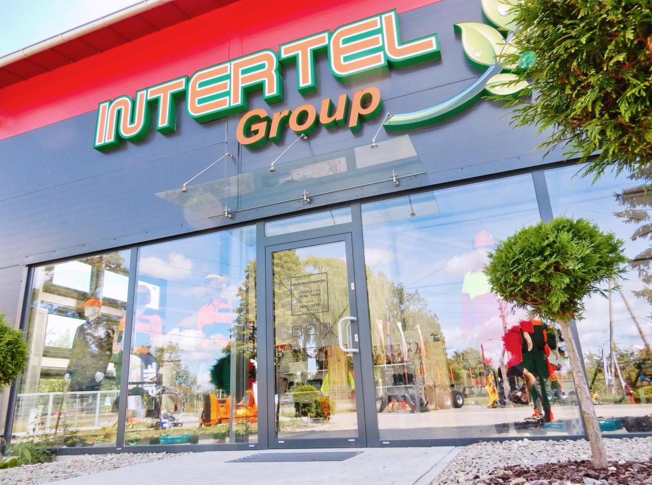 Fasada budynku firmy Intertel Group z dużymi oknami wystawowymi, na których widoczne są manekiny w strojach roboczych i sprzęt ogrodniczy.