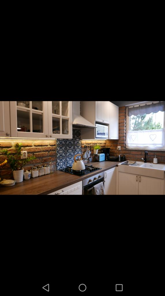 Kuchnia w stylu rustykalnym z białymi szafkami, drewnianym blatem, ceglaną ścianą i dekoracyjnymi płytkami nad kuchenką. Na blacie czajnik, przyprawy i roślina w doniczce.