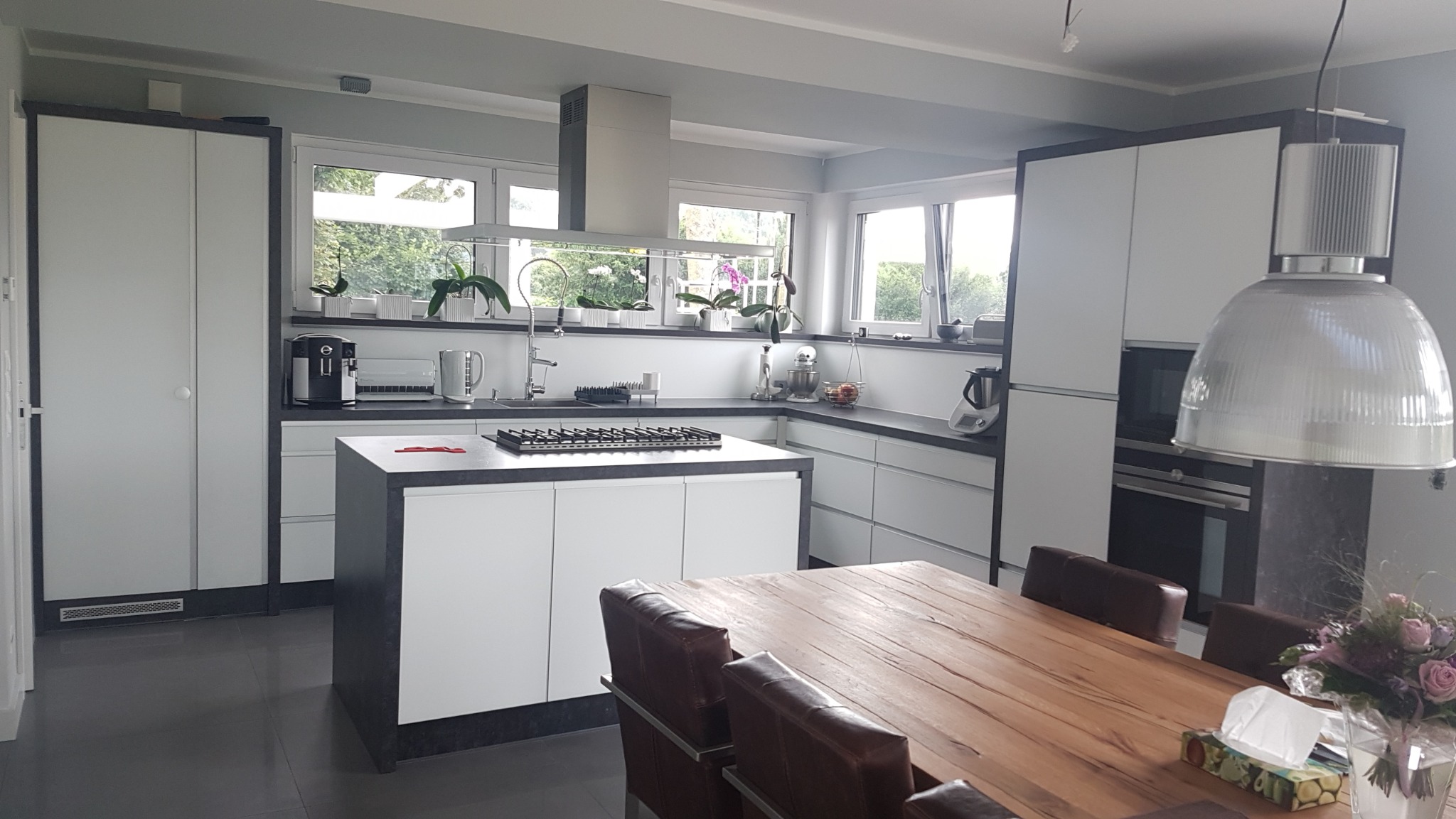 Nowoczesna kuchnia z białymi szafkami, szarym blatem i wyspą kuchenną z płytą grzewczą, widok na stół jadalny z krzesłami i bukietem kwiatów, oświetlenie lampą wiszącą.