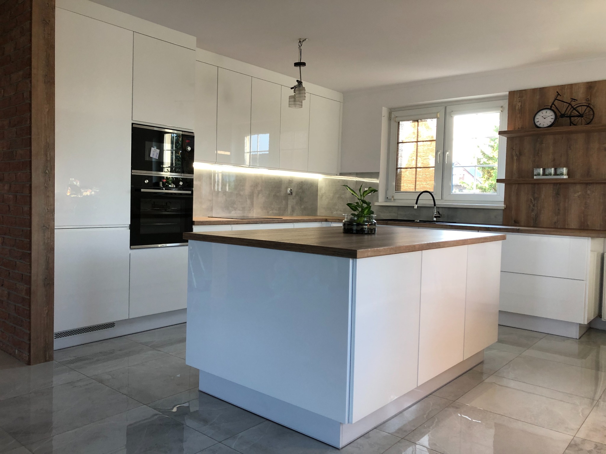 Biała kuchnia z wyspą, połyskującymi frontami szafek, drewnianym blatem, ceglanym elementem ściany i dekoracyjnym zegarem w kształcie roweru na półce.