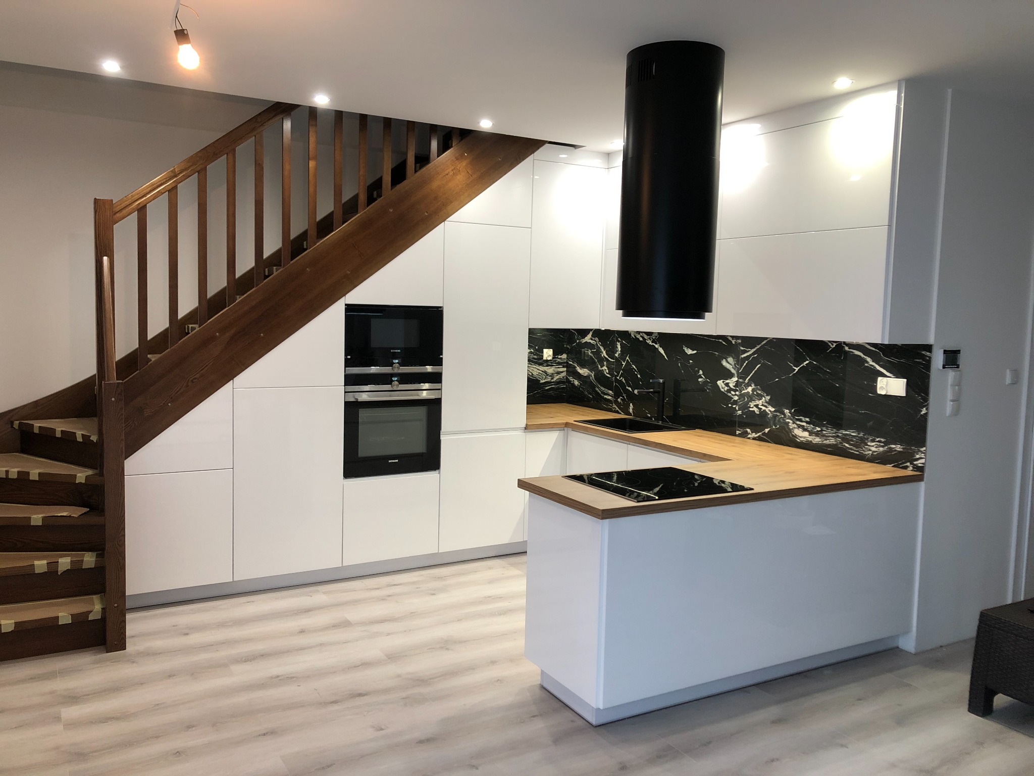 Nowoczesna biała kuchnia z drewnianym blatem i czarnym panelem nad blatem, drewniane schody prowadzą na górę, widoczny czarny okrągły okap.