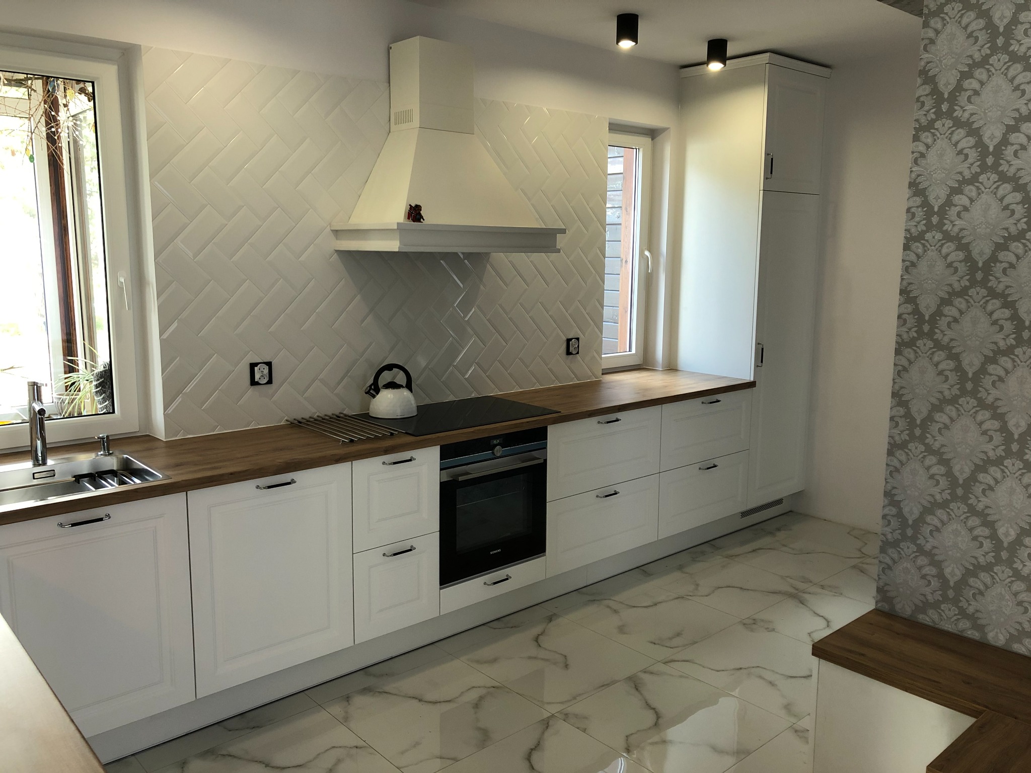 Nowoczesna, biała kuchnia z drewnianym blatem i białymi płytkami ułożonymi w jodełkę na ścianie, z czajnikiem na płycie grzewczej.