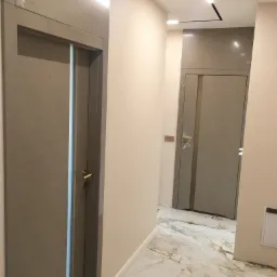 Wnętrze korytarza z marmurową podłogą, beżowymi ścianami i dwiema szarymi drzwiami z delikatnym wzorem, oświetlonego nowoczesnym, liniowym oświetleniem sufitowym.