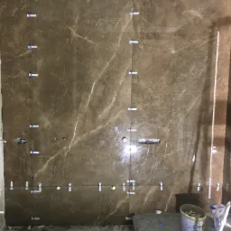Ściana w trakcie układania brązowych płytek imitujących marmur, z widocznymi otworami na instalacje i systemem poziomowania płytek, surowy stan posadzki, kabel wiszący z sufitu.