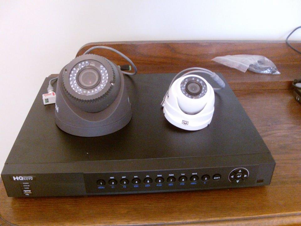 Dwie kamery kopułkowe, szara i biała, leżące na czarnym rejestratorze DVR na drewnianej powierzchni.