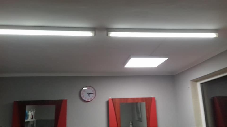 Wnętrze salonu fryzjerskiego z trzema lampami sufitowymi: dwie liniowe świetlówki i jedna kwadratowa LED, oraz dwa lustra w czerwonych ramach i okrągły zegar ścienny.