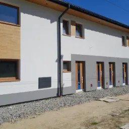 Nowoczesny budynek z białą elewacją, drewnianymi akcentami okiennymi i szarymi wnękami drzwiowymi, widok z zewnątrz na teren budowy z gruzem i paletami.