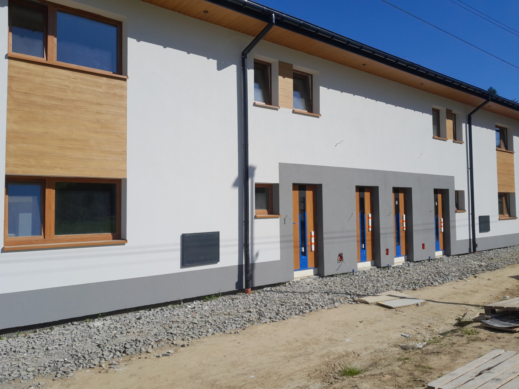 Nowoczesny budynek z białą elewacją, drewnianymi akcentami okiennymi i szarymi wnękami drzwiowymi, widok z zewnątrz na teren budowy z gruzem i paletami.