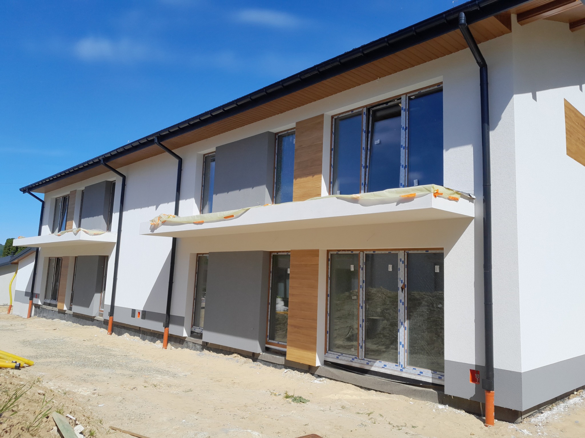 Nowoczesny szeregowiec z biało-szarą elewacją i elementami drewnopodobnymi, widoczne zabezpieczenia okien folią, teren wokół budynku w trakcie prac wykończeniowych.