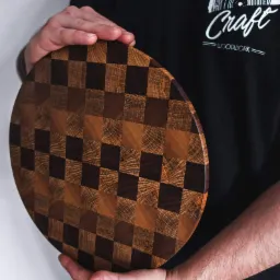 Okrągła deska do krojenia z drewna w szachownicę, trzymana przez mężczyznę w czarnej koszulce z logo 'Craft Woodwork'.