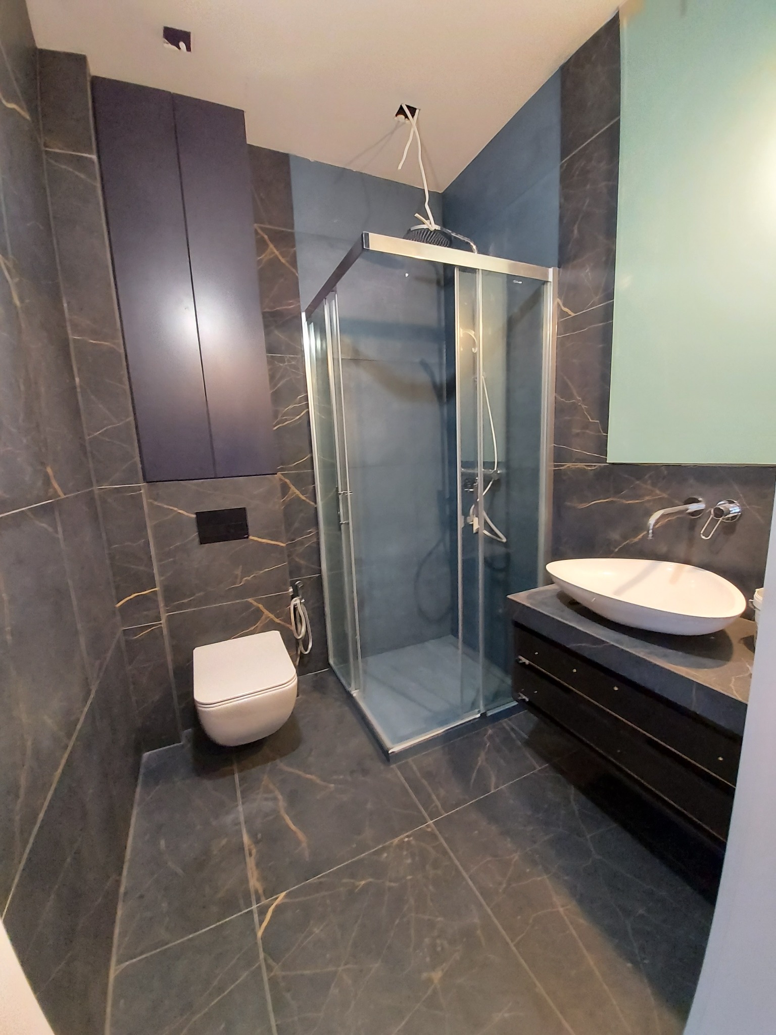 Nowoczesna łazienka z grafitowymi płytkami imitującymi marmur, kabiną prysznicową ze srebrnymi profilami, podwieszaną toaletą i umywalką nablatową w kształcie łzy.