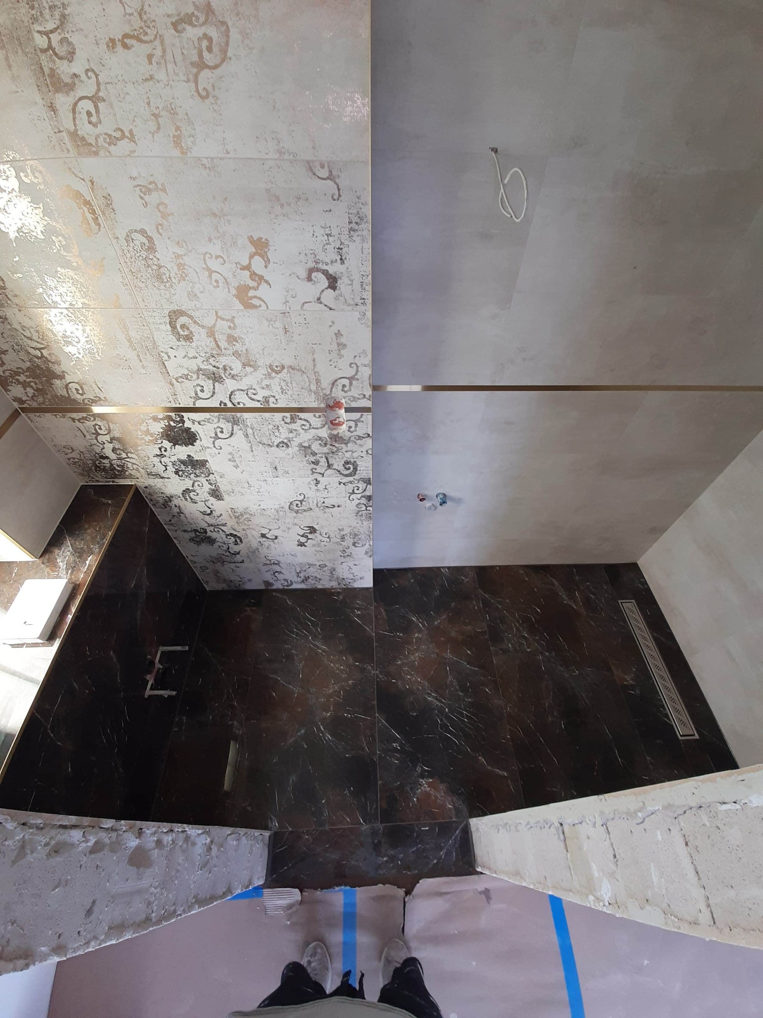 Łazienka w trakcie wykańczania: połowa ścian wyłożona wzorzystymi, beżowo-złotymi płytkami, druga połowa gładkimi, szarymi płytkami. Na podłodze ciemne, marmurkowe płytki. Widoczne niedokończone...