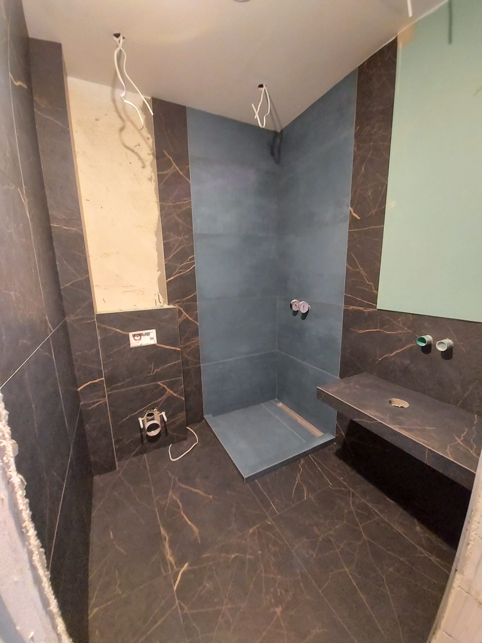 Wnętrze łazienki w trakcie prac wykończeniowych: ciemne płytki imitujące marmur na podłodze i ścianach, kabina prysznicowa typu walk-in z odpływem liniowym, częściowo wykończona ściana z instalacją...