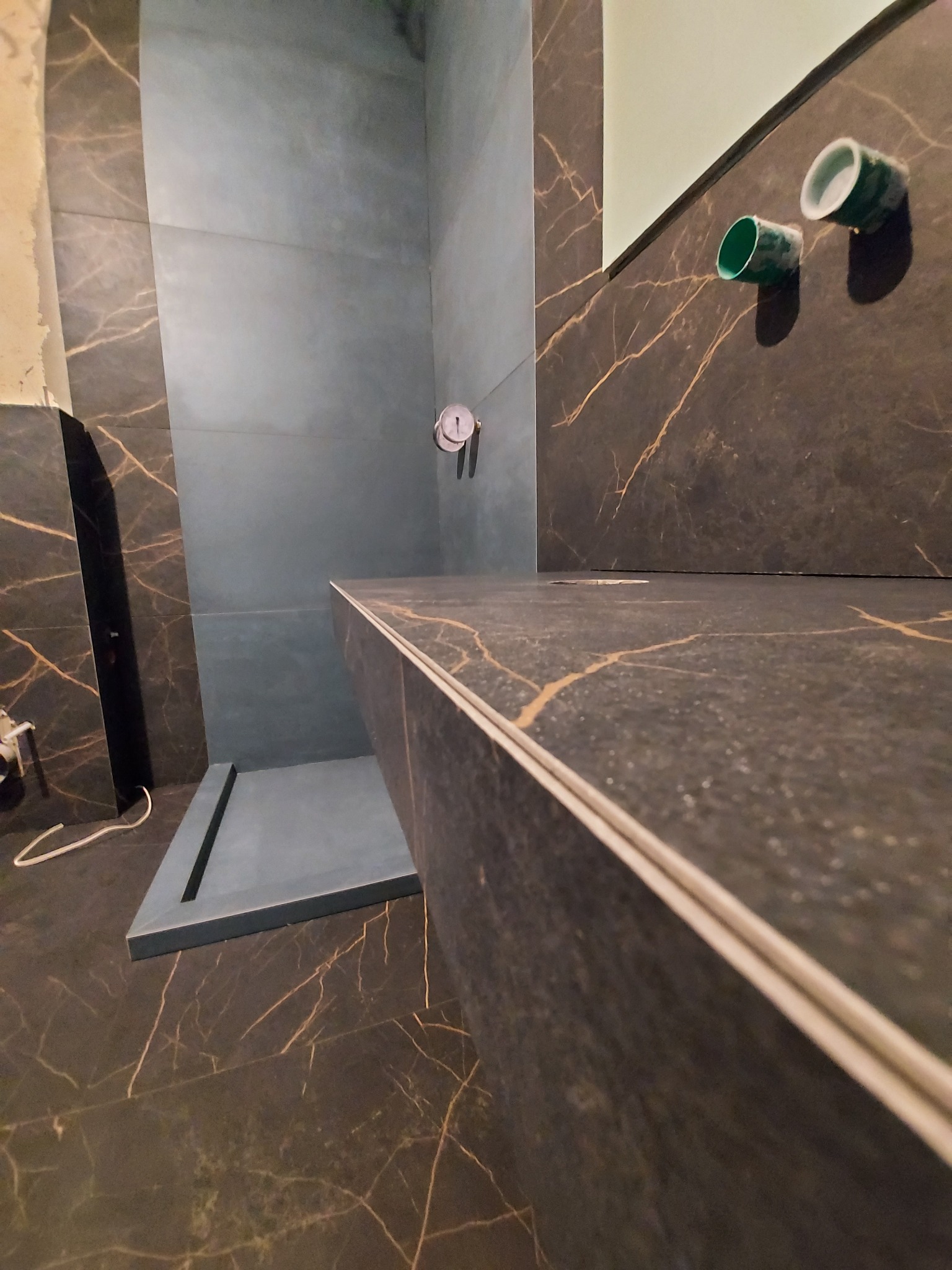 Niedokończona kabina prysznicowa z szarymi i brązowymi płytkami imitującymi marmur, widoczne rury instalacji wodnej i odpływ liniowy w posadzce.