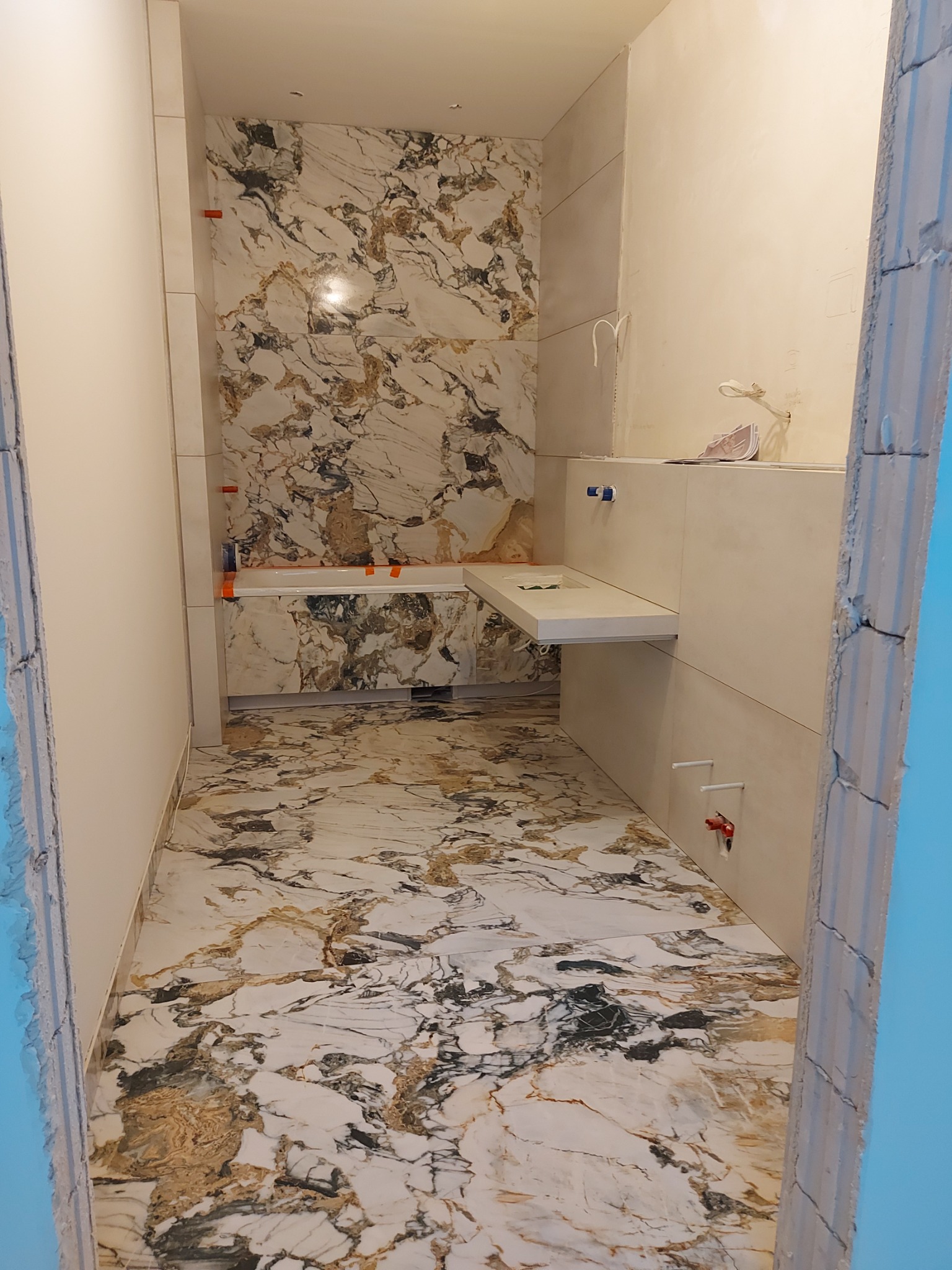 Łazienka w trakcie wykańczania z marmurową podłogą i ścianami, widoczne fragmenty instalacji wodnej i sanitariatów, surowy stan ościeżnicy po lewej stronie.