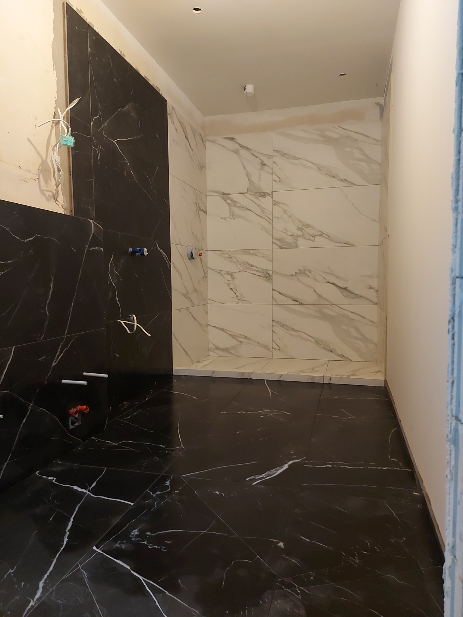 Łazienka w trakcie wykończenia w Ełku: położone czarno-białe płytki imitujące marmur na podłodze i ścianach, widoczne wystające kable i rury przed montażem armatury.