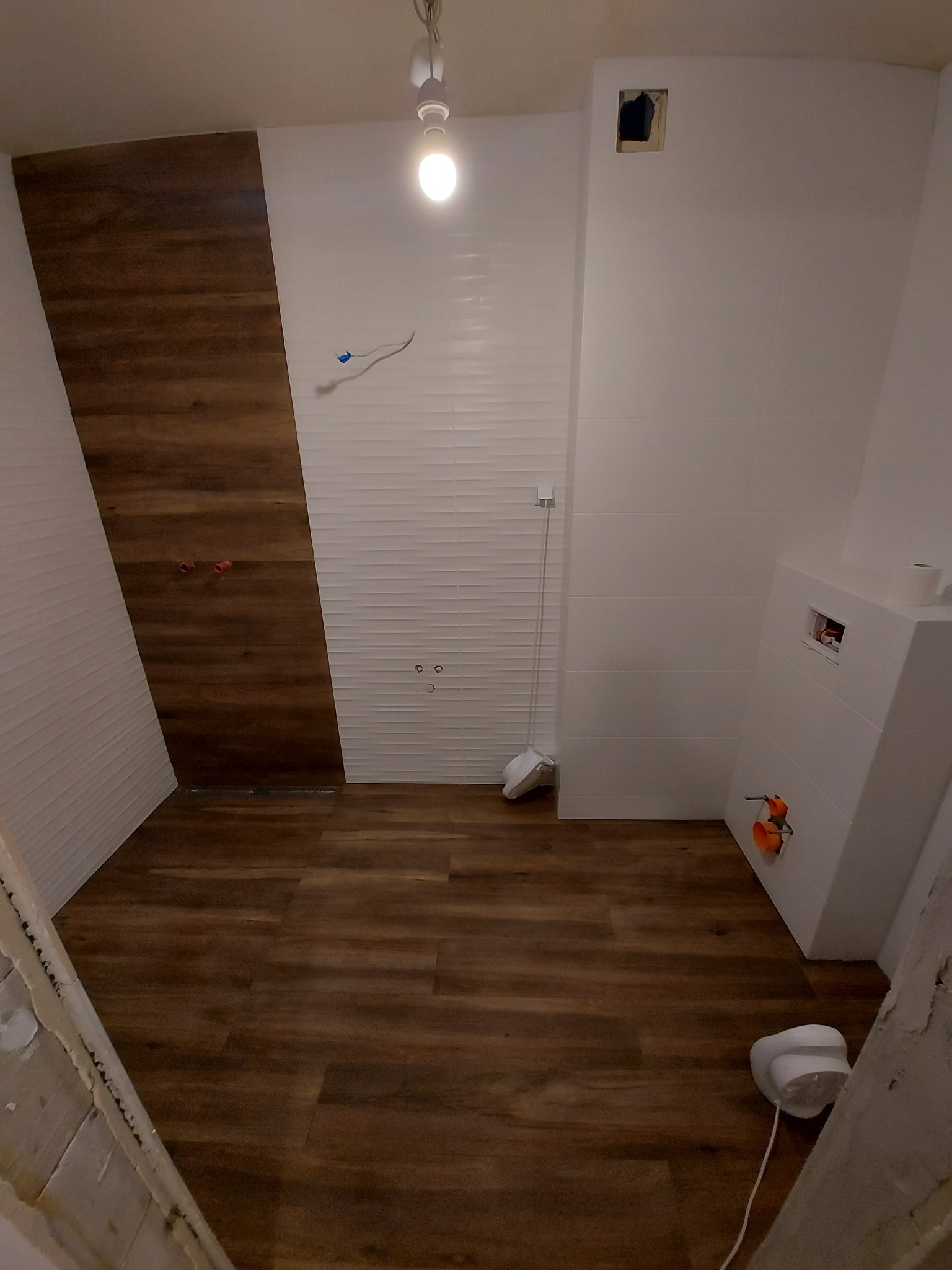 Łazienka w trakcie remontu: drewnopodobne panele ścienne i podłogowe, białe płytki ścienne z poziomym wzorem, zabudowa WC z widocznymi elementami instalacji, surowy narożnik ściany z resztkami farby.