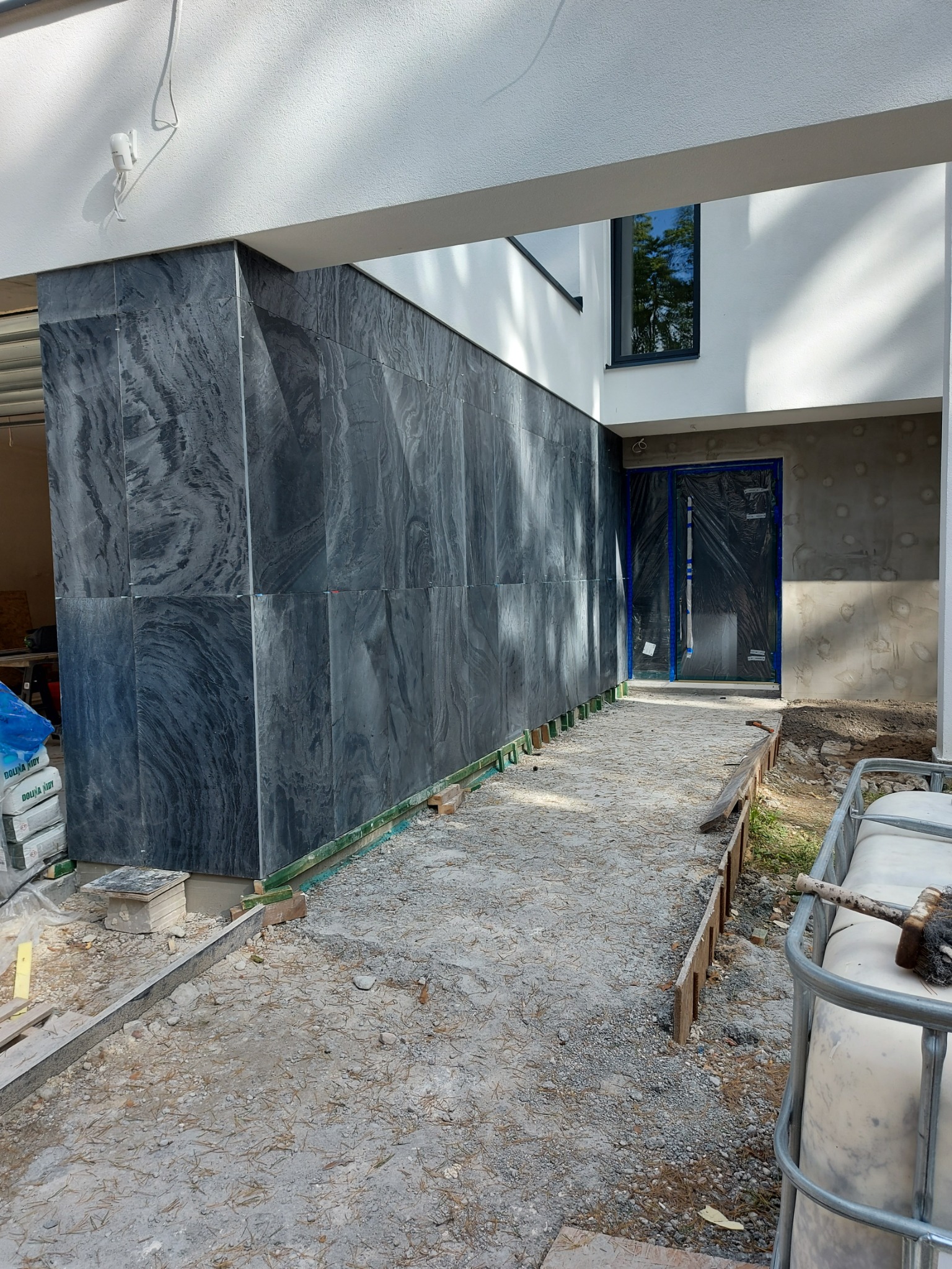 Elewacja budynku w trakcie wykończenia, z widocznymi płytami z szarego kamienia o wyraźnym wzorze żyłkowania, zabezpieczone drzwi wejściowe folią i drewnianymi elementami szalunku wzdłuż ścieżki.