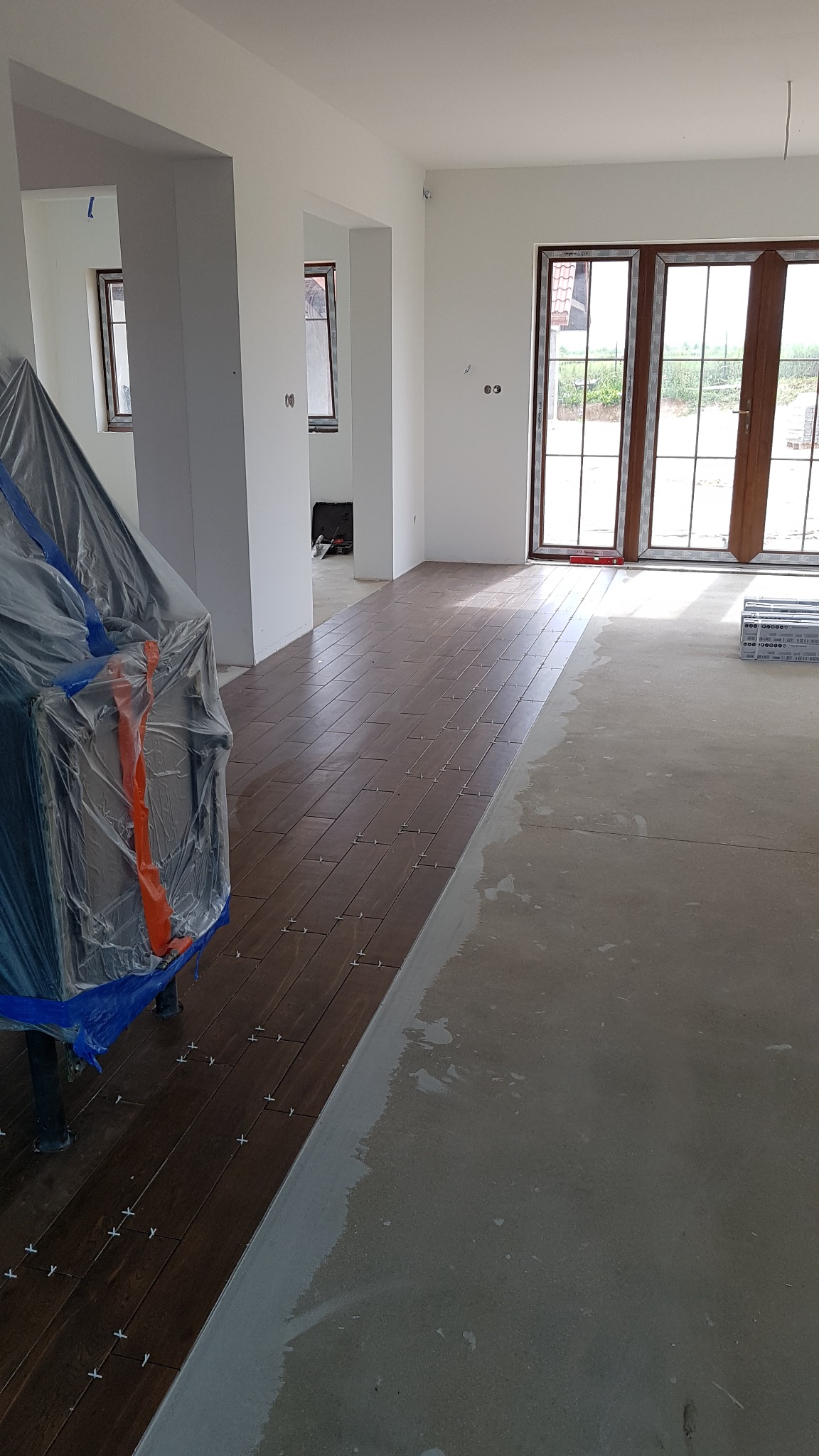 Układanie brązowych płytek podłogowych z krzyżykami dystansowymi w przestronnym, jasnym pomieszczeniu, widoczny fragment podłogi przygotowanej pod wylewkę.