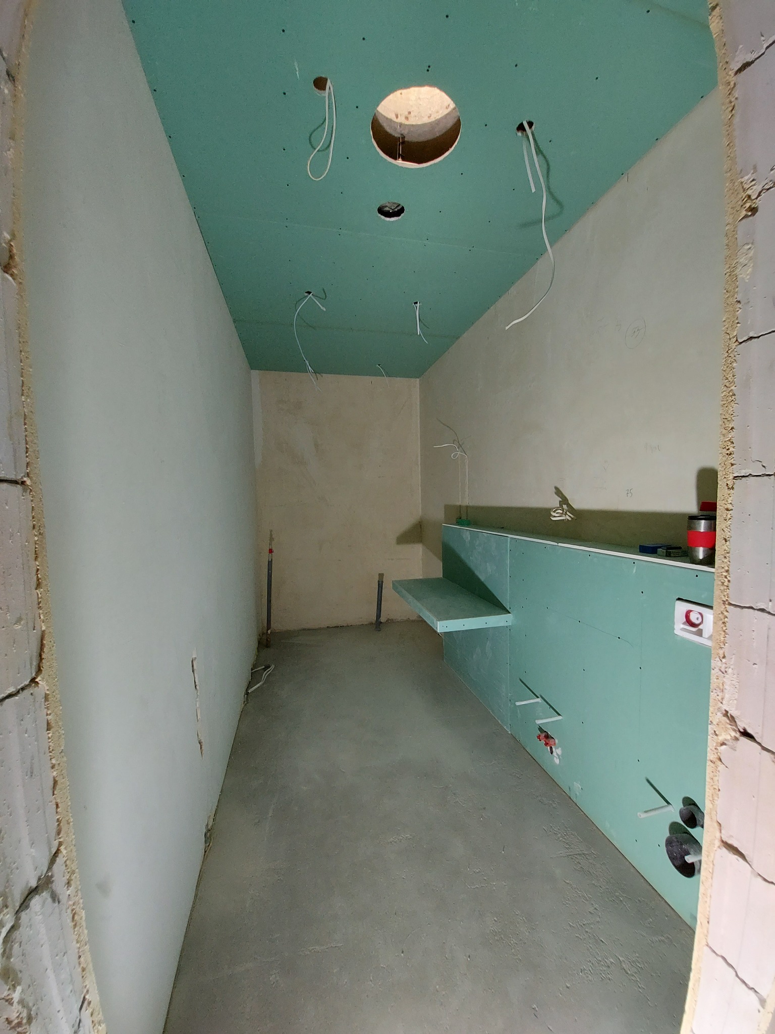 Łazienka w trakcie wykańczania: zielone płyty gipsowo-kartonowe na ścianach i suficie, widoczne instalacje hydrauliczne i elektryczne, surowy beton na podłodze.