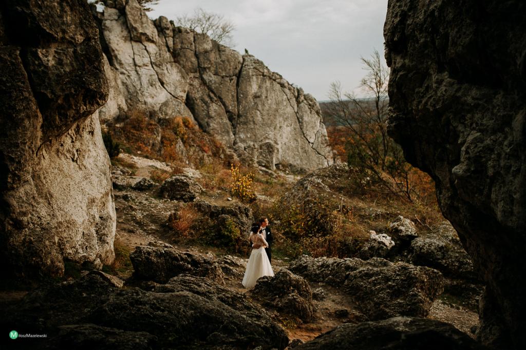Para młoda w strojach ślubnych pozuje wśród wapiennych skał na tle jesiennego krajobrazu.