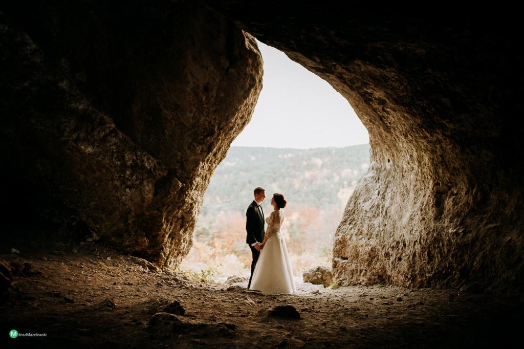 Para młoda w jaskini, panna młoda w białej sukni z koronkowymi rękawami, pan młody w czarnym smokingu, w tle wzgórza z drzewami o jesiennych barwach.