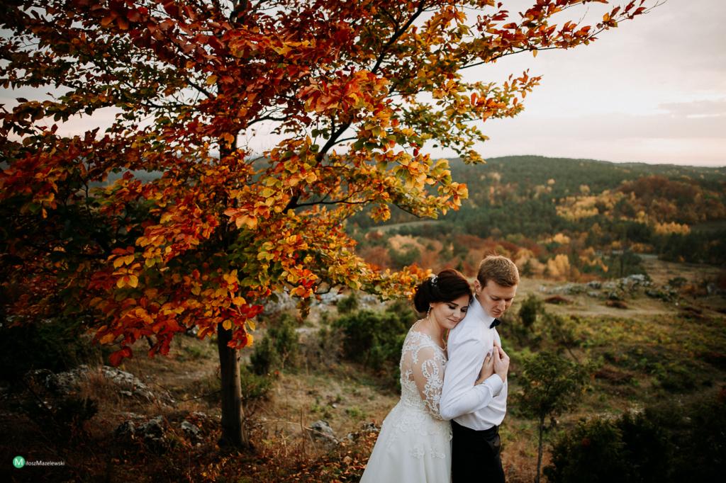Para młoda obejmująca się pod drzewem w jesiennych barwach na tle zalesionych wzgórz