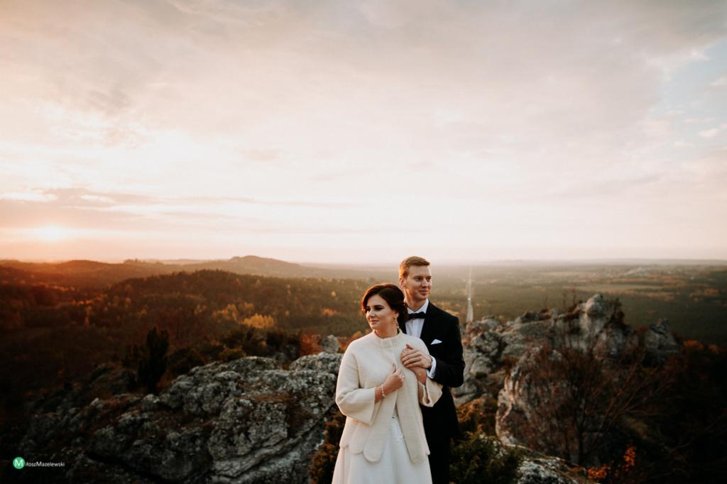 Para młoda w strojach ślubnych stoi na skalistym wzgórzu o zachodzie słońca, z widokiem na zalesione tereny i odległe miasto.