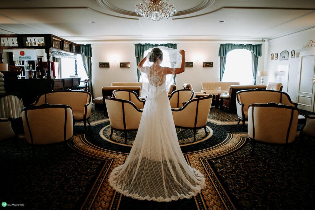 Panna młoda w białej sukni ślubnej, stojąca tyłem, trzymająca welon w luksusowym wnętrzu hotelowym z kremowymi fotelami i wzorzystym dywanem.