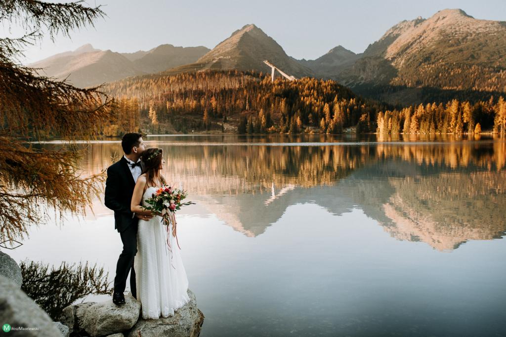 Para młoda w Częstochowie pozuje na tle górskiego jeziora i jesiennego lasu, odbijających się w tafli wody, panna młoda z bukietem w ręku.