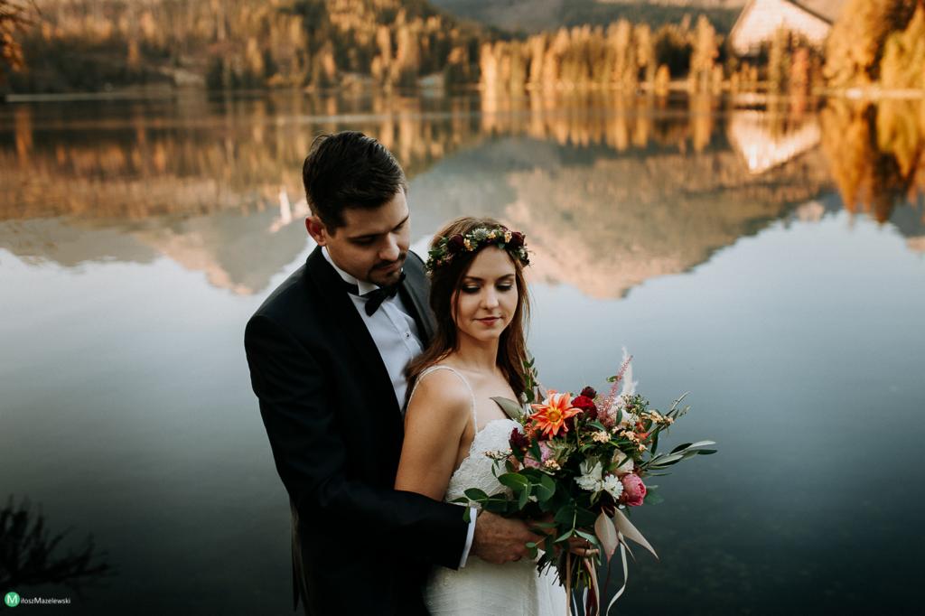 Para młoda w eleganckich strojach pozuje na tle jeziora z odbijającymi się górami, panna młoda trzyma bukiet z pomarańczowymi daliami i ma wianek na głowie.