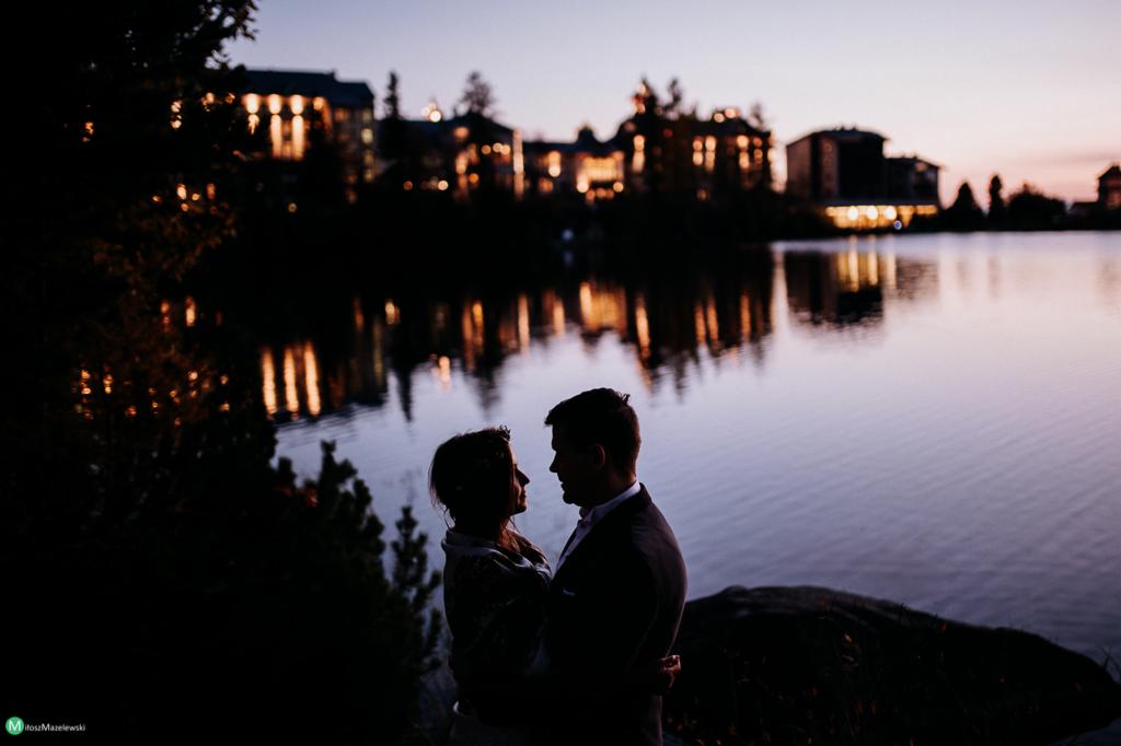 Silhouettes of embracing couple by a lake at dusk, with building lights reflecting on the water.