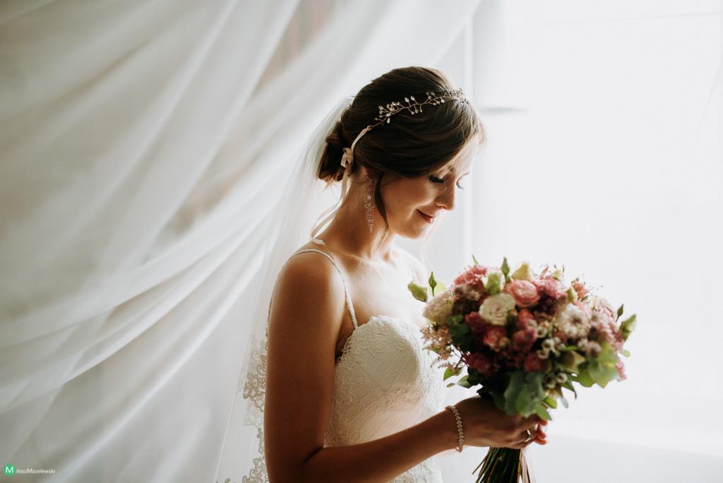 Panna młoda w koronkowej sukni, z delikatnym bukietem różowych i białych kwiatów, spogląda w dół, z tiulem powiewającym za nią, w tle jasne okno.